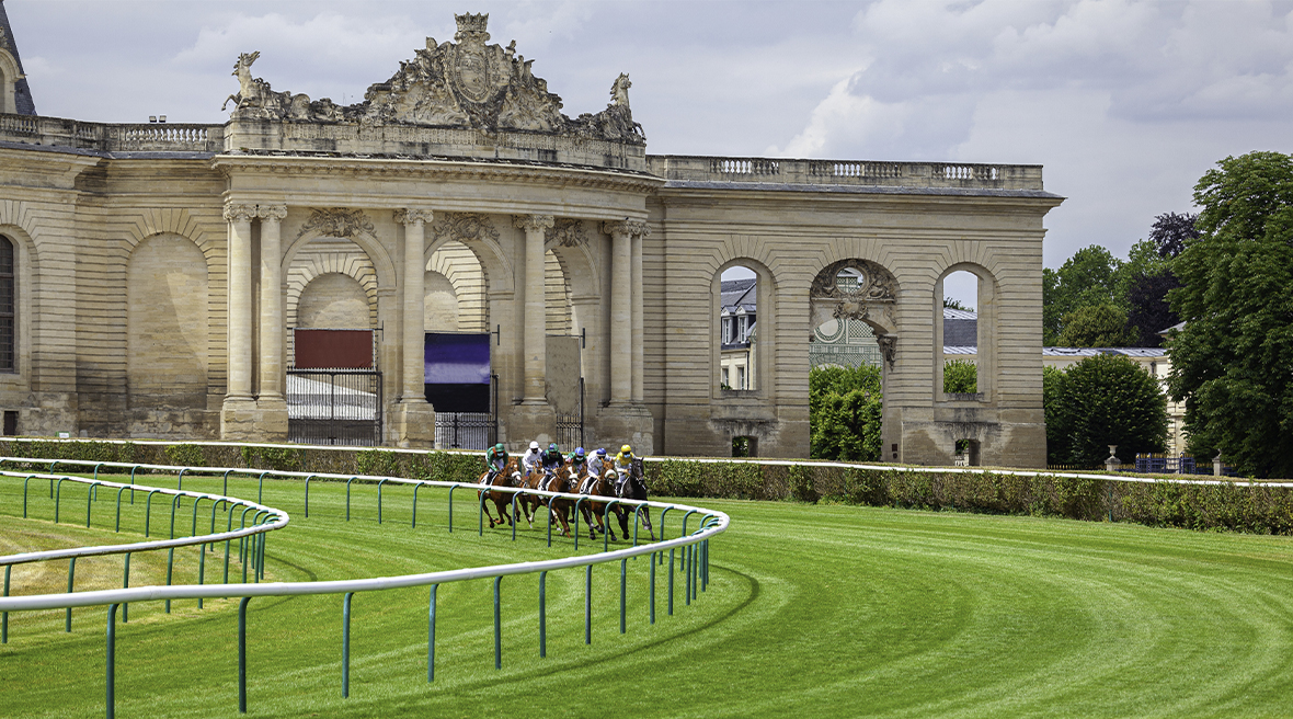 a pale-stone palatial arch and low building alongside, with horse racing track in the foreground